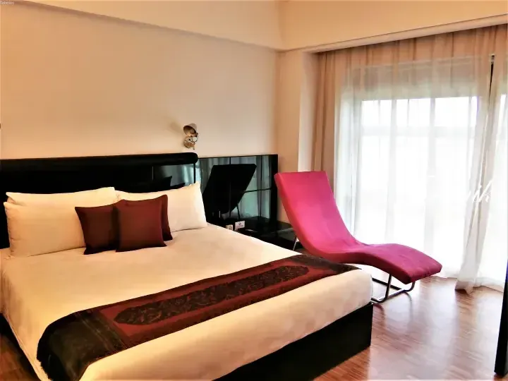 Modern hotel bedroom with a king bed, pink chaise lounge, and large window with sheer curtains