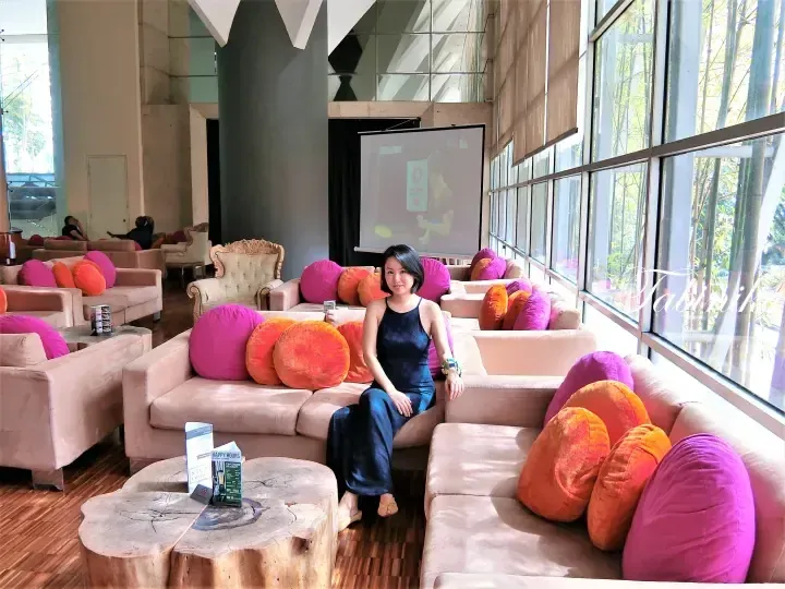 Woman sitting on a sofa in a lounge with colorful cushions and large windows at a modern property.