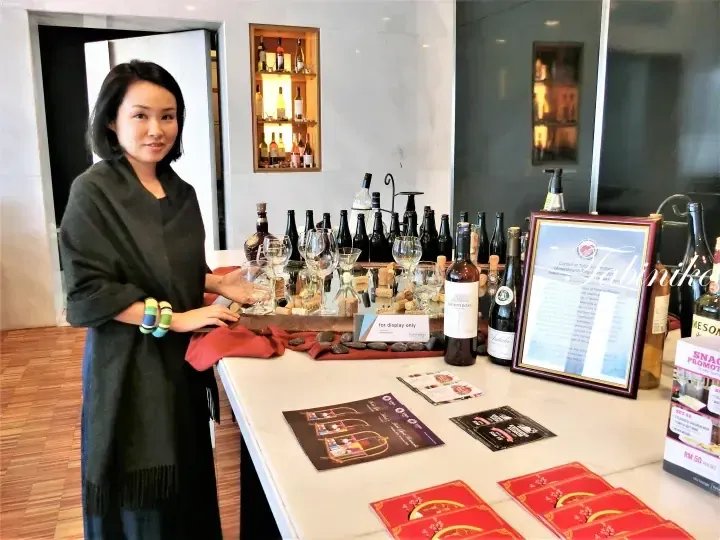 Woman presenting wine bottles and tasting glasses at a display table in Hotel Maya, featuring promotional materials and a framed notice.