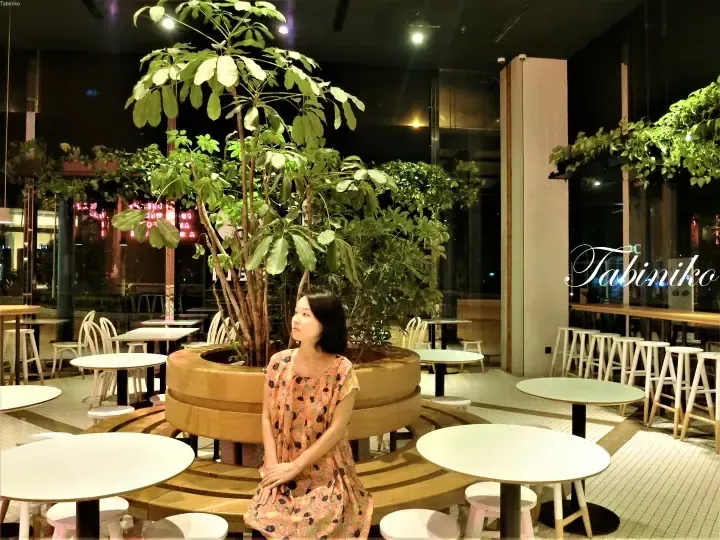 Woman in floral dress sitting on curved wooden bench surrounded by lush plants in modern cafe interior.