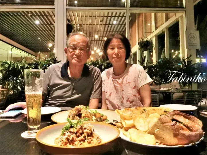 An older couple dining at an outdoor restaurant with plates of pasta, pizza, and a Tiger beer glass.