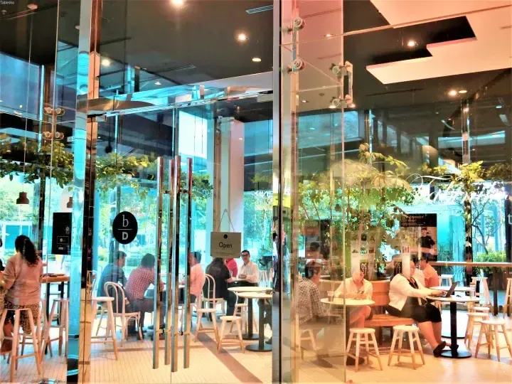 Modern cafe interior with glass walls, wooden furniture, and patrons dining, featuring a black b/D logo on the window.