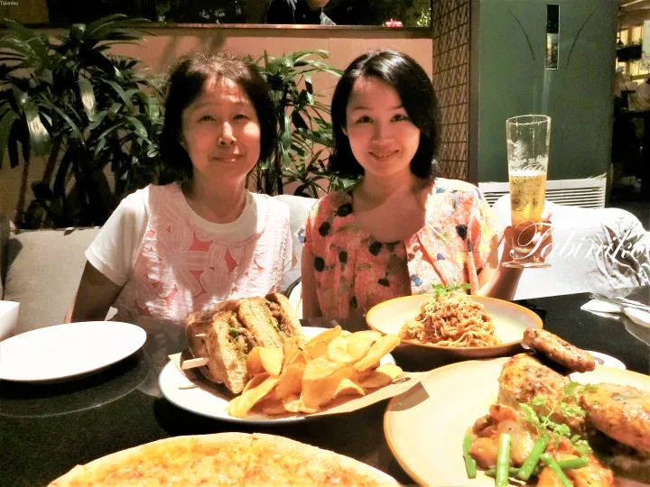 サンドイッチやパスタ、チキンウィングが並んだテーブルで笑顔を見せる二人の女性。