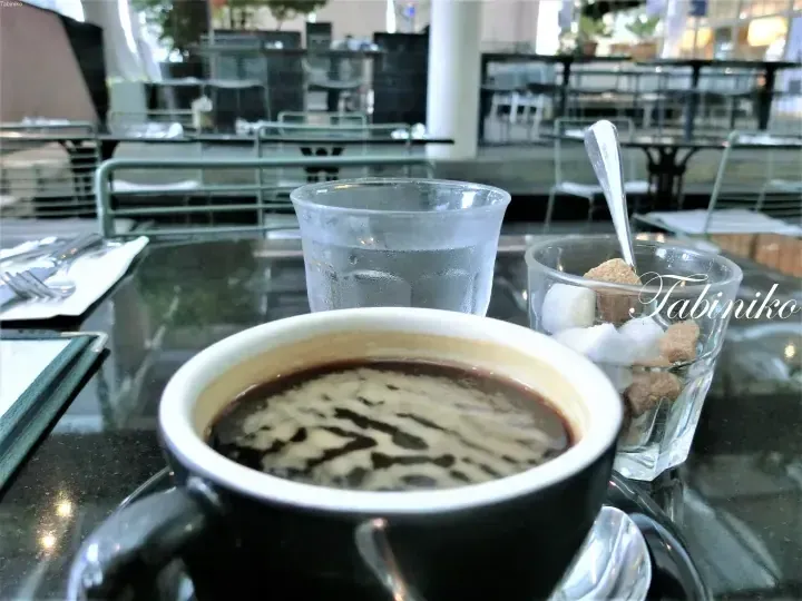 A cup of coffee with milk swirls sits on a table alongside a glass of water and sugar cubes in a bright cafe setting.