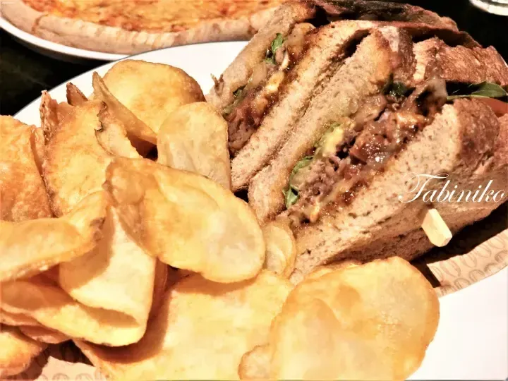 A hearty sandwich with meat and greens served alongside crispy potato chips on a white plate.