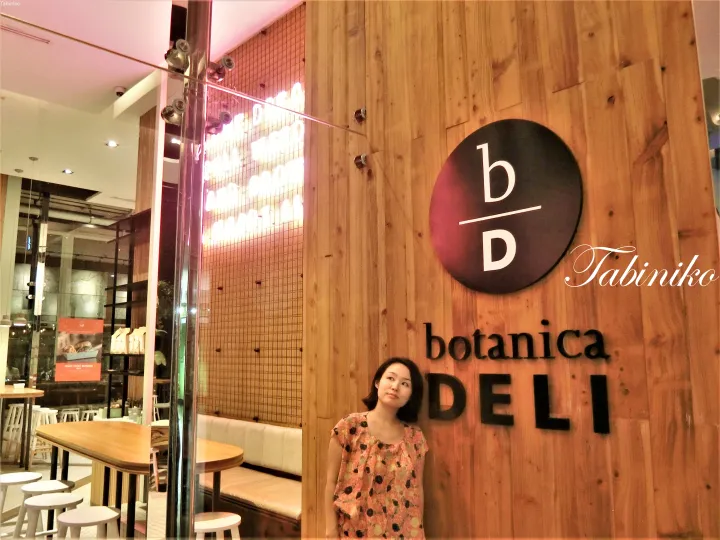 木目調の壁と「b/D」ロゴが目立つBotanica Deliの外でポーズをとる女性。
