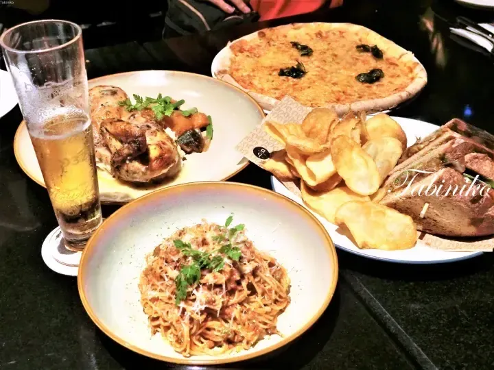 Table spread featuring pizza, pasta, chicken, sandwich, chips, and beer at a dining setting