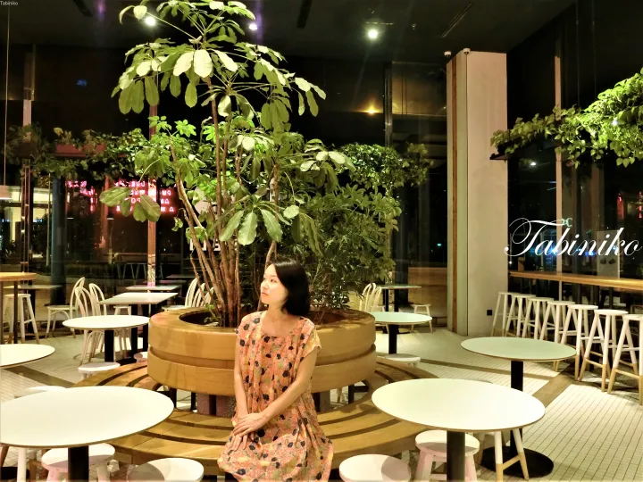 緑豊かな植物に囲まれたモダンなカフェの曲線木製ベンチに座る花柄ドレスの女性。