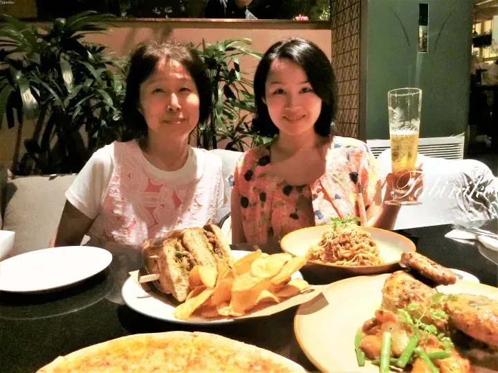 Two women smile at a dining table filled with plates of sandwiches, pasta, and chicken wings.