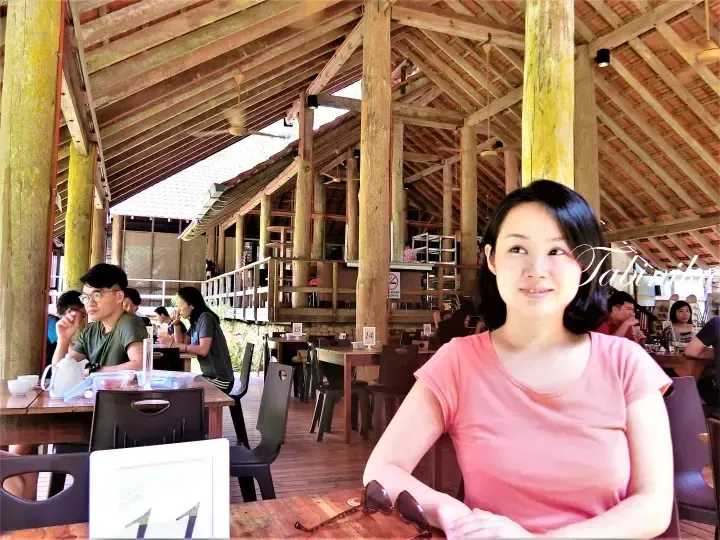 A woman in a pink shirt sits at a table in a spacious wooden open-air restaurant with high ceilings and other diners.