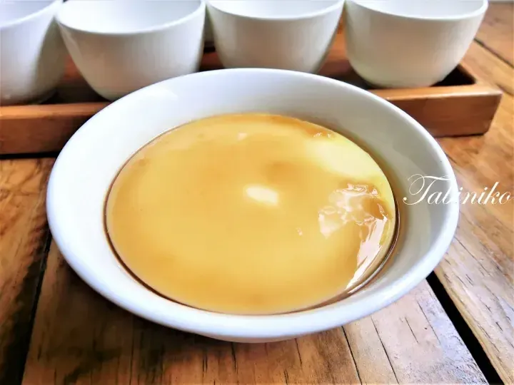 A smooth yellow custard dessert in a white bowl with caramel sauce, served on a wooden table.