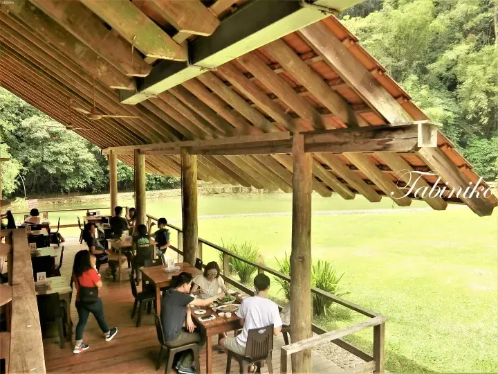 Diners enjoy a meal at an open-air wooden restaurant deck overlooking a green lake and lush forest.