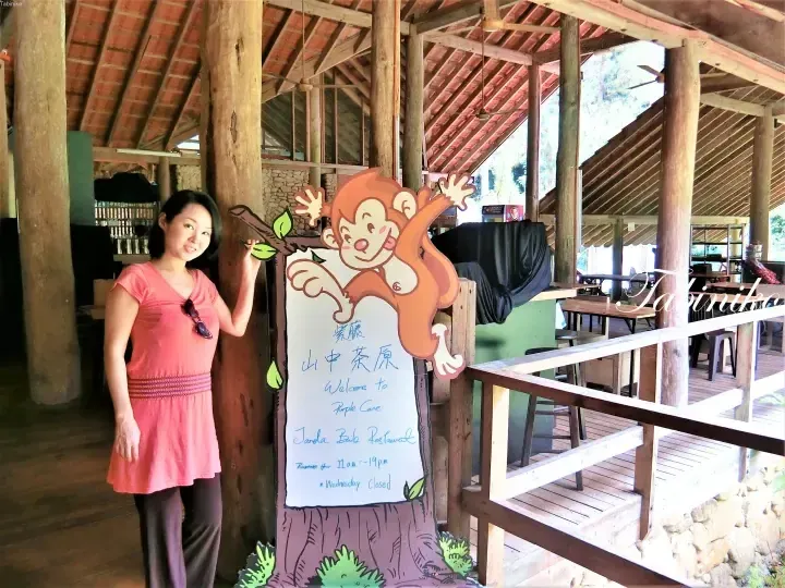 Woman posing beside a sign for Janda Baik Restaurant at Purple Cane, featuring a cartoon monkey and handwritten welcome message.