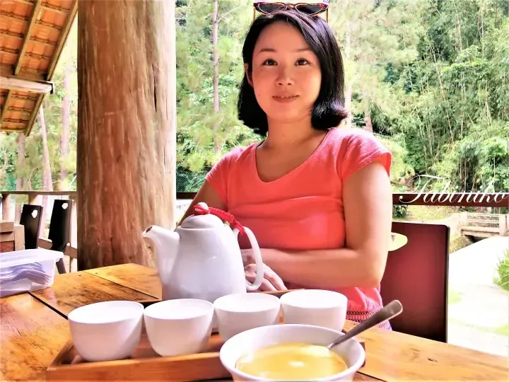 Woman in pink shirt enjoying tea and dessert at a wooden table with a scenic forest view.