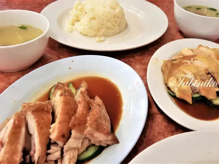 Slices of Hainanese chicken rice with dark sauce and cucumber on a plate, accompanied by a mound of fragrant rice and bowls of soup.