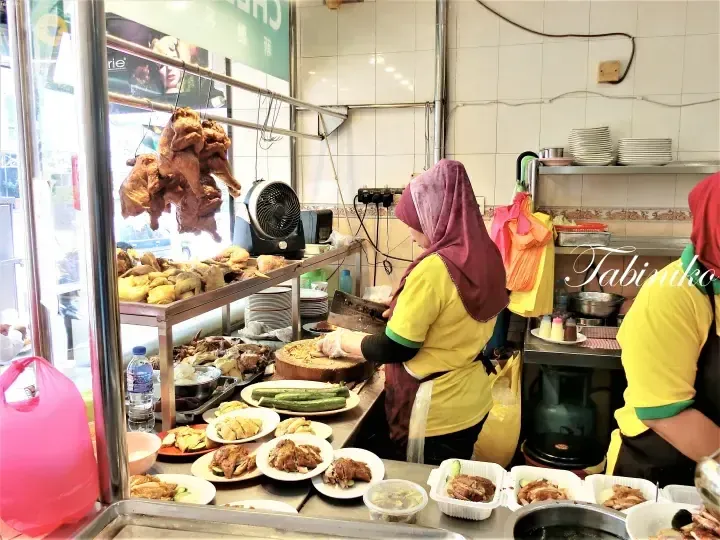 A vendor chops roasted chicken at a busy food stall with hanging roasted ducks and plates of prepared dishes.
