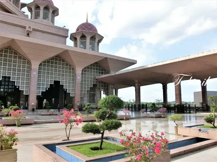 Pink Putra Mosque exterior with geometric windows, manicured gardens, and reflecting pool under a blue sky.