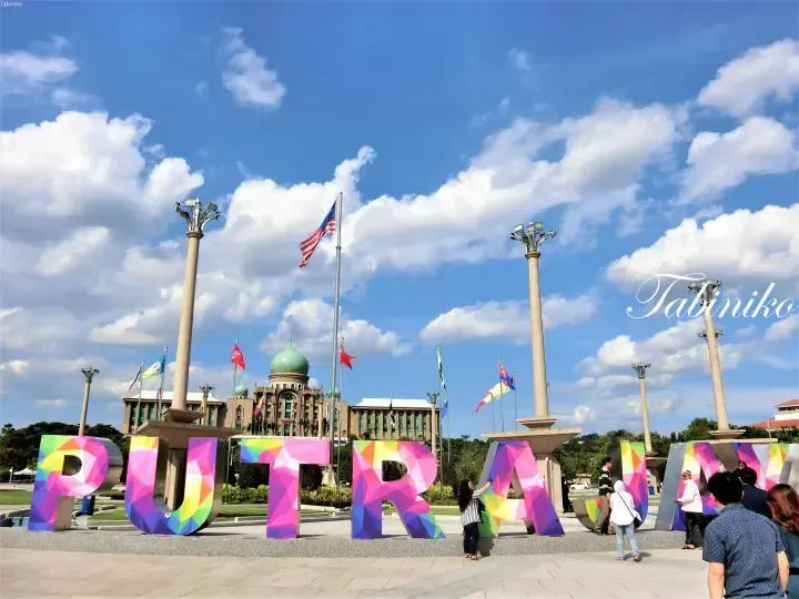 Colorful PUTRA letters in front of the Perdana Putra building with Malaysian flags under a blue sky.