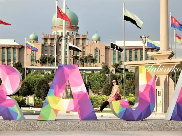 A woman sits on a colorful geometric letter sculpture in front of the iconic green-domed Putra Mosque in Putrajaya.