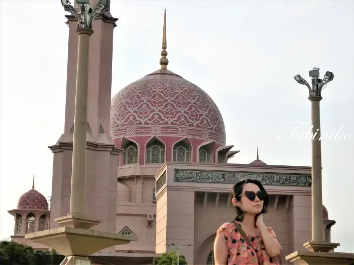 Woman wearing sunglasses stands in front of the iconic pink dome and minaret of Putra Mosque in Putrajaya.