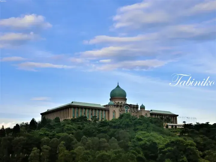The majestic Putra Mosque with its iconic green dome sits atop a lush hill under a bright blue sky.