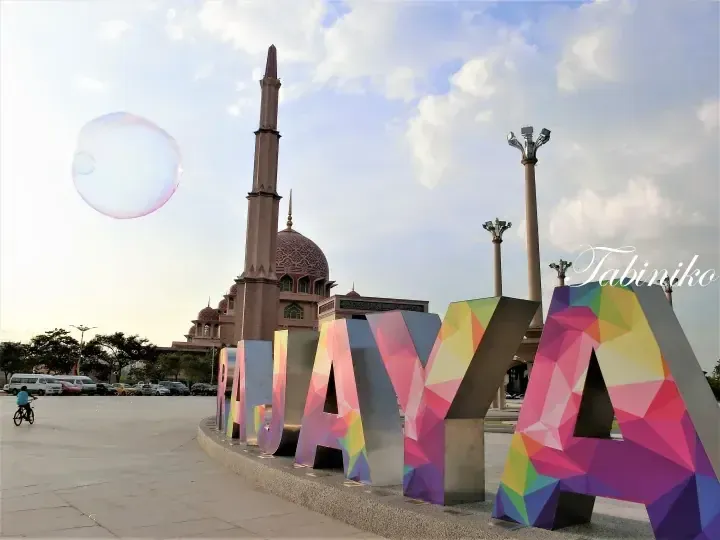 Colorful geometric I Love KL sign in front of the pink Putra Mosque under a blue sky with a large soap bubble floating nearby.