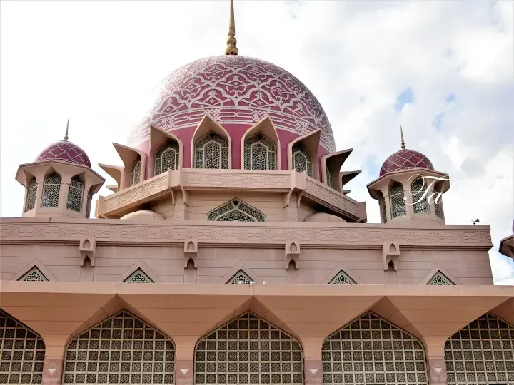 Pink dome with intricate white geometric patterns and arched windows of a mosque under a cloudy sky