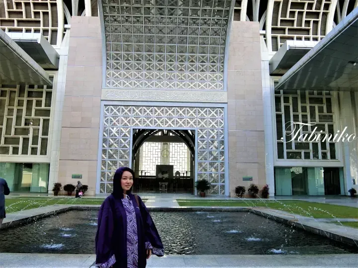 Woman in purple attire stands before the grand entrance of the National Mosque Malaysia, featuring intricate geometric patterns and a fountain.