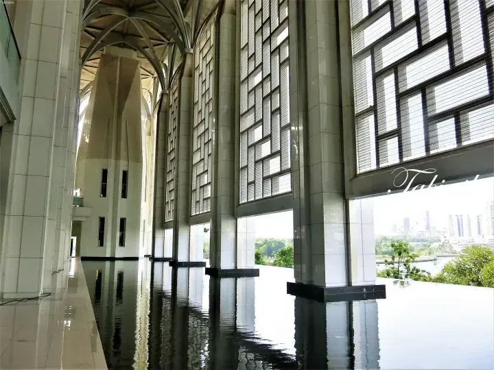 Interior view of the Islamic Arts Museum Malaysia featuring tall white pillars, a reflective water floor, and large windows overlooking the city.