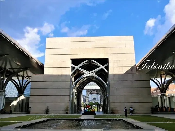 Architectural entrance with pointed arches framing the National Mosque in Kuala Lumpur under a blue sky.