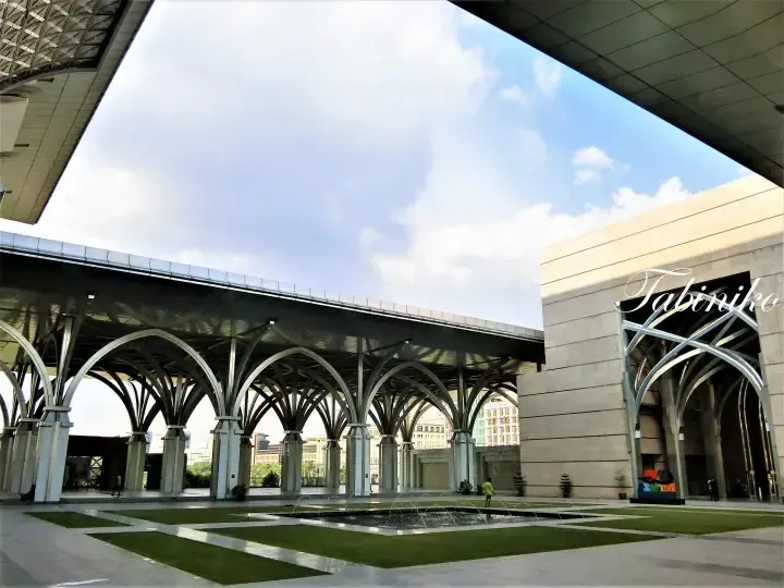 Architectural colonnade with arched steel beams at Suria KLCC, featuring a central water fountain and manicured green lawn under a bright sky.
