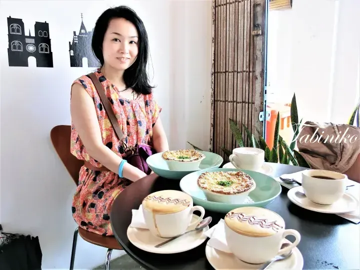 Woman smiling at a cafe table with coffee and baked dishes, featuring a wall decal of a European-style building.