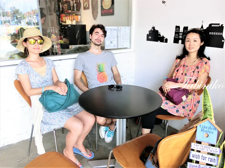 パイナップルTシャツの男性と2人の女性がカフェの屋外席で丸テーブルを囲んで座っている。