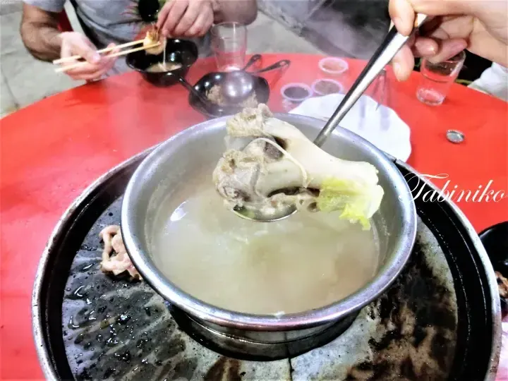 A large bone in milky soup is lifted with a ladle from a traditional Malaysian charcoal hotpot on a red table.