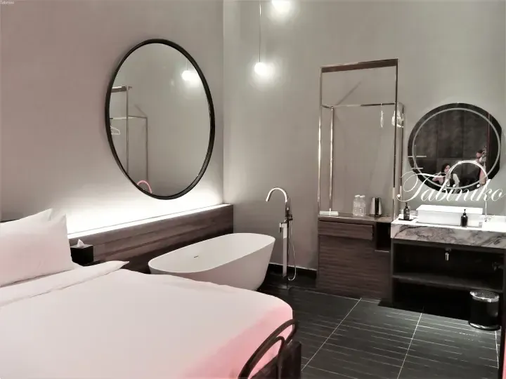 Modern hotel room with a freestanding bathtub, large round mirror, and marble vanity.