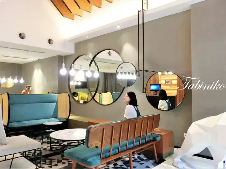 Modern hotel lobby with blue velvet seating, geometric mirrors, and a woman sitting on a wooden bench.
