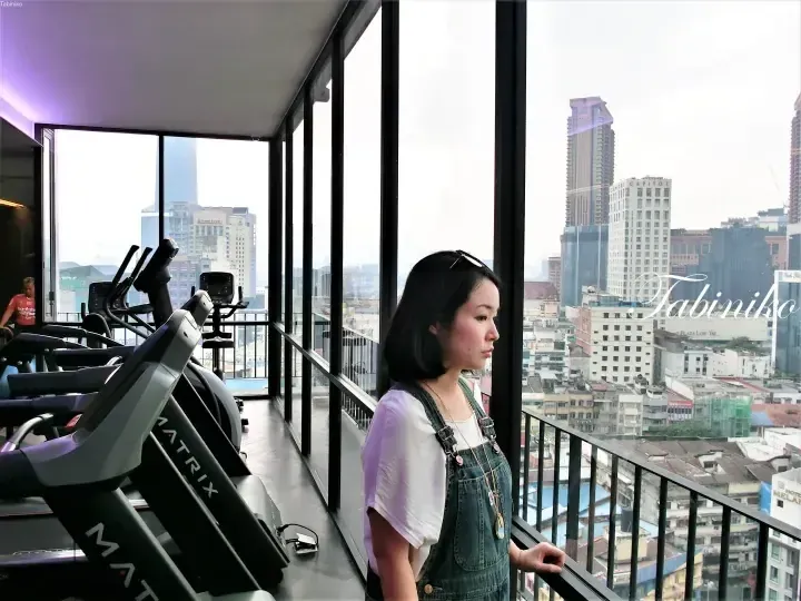 Woman in denim overalls looking out gym window at Kuala Lumpur skyline featuring Capitol Hotel.