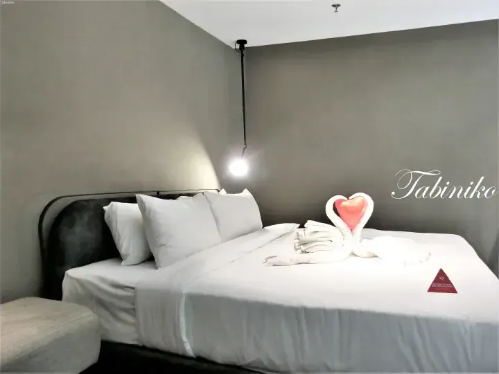 Modern hotel room with grey walls, white bedding, a towel swan with a heart, and a hanging light fixture.