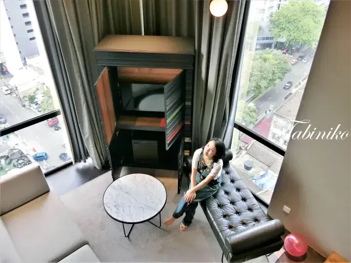 Woman sitting on a black leather bench in a modern hotel room with a marble coffee table and city view.