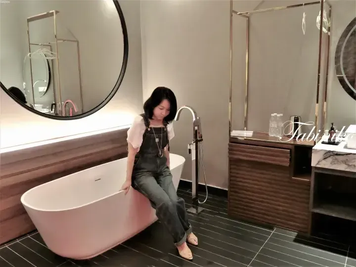 Woman sitting on a freestanding bathtub in a modern hotel bathroom with dark tiled flooring and a large round mirror.