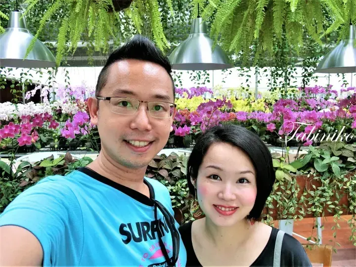 A smiling couple takes a selfie in a vibrant greenhouse surrounded by colorful orchids and hanging ferns.