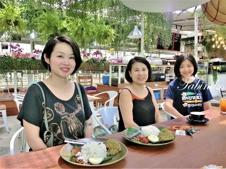 Three women dining at a lush green cafe, enjoying plates of rice and curry in a vibrant garden setting.
