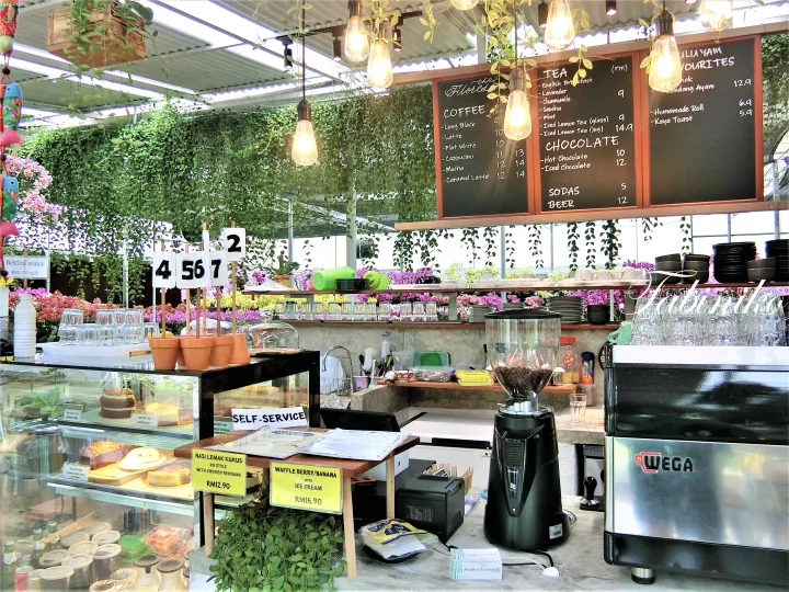 エスプレッソマシンとメニューボード、緑の植物が飾られたカフェのカウンター。背景には蘭の花が並ぶ。