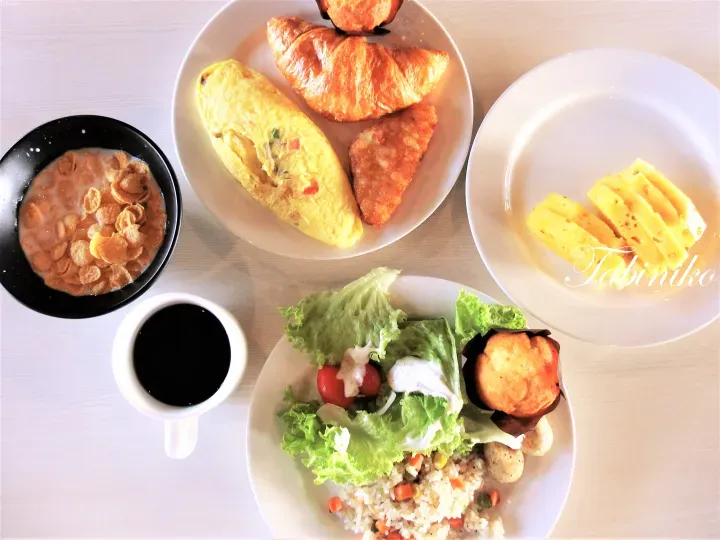 A breakfast spread featuring an omelette, croissant, cornflakes, pineapple slices, salad, and coffee.