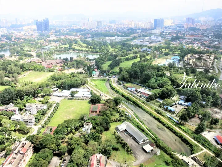 Aerial view of a lush green landscape with a river, lake, and golf course, overlooking a city skyline with high-rise buildings in the distance.