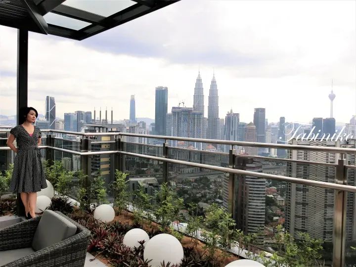 Woman in polka-dot dress stands on a balcony overlooking the Petronas Twin Towers and Kuala Lumpur skyline.