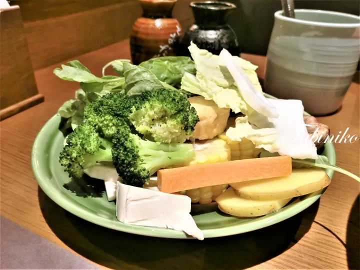 A green plate filled with fresh vegetables including broccoli, corn, carrots, potatoes, and leafy greens.