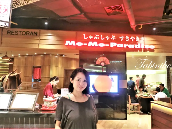 A woman stands smiling in front of the Mo-Mo-Paradise shabu shabu and sukiyaki restaurant entrance.
