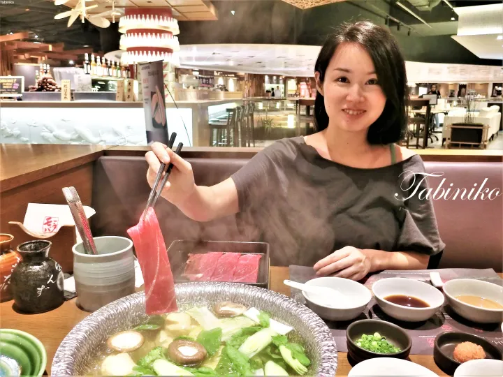 A woman dining at a restaurant holds chopsticks with a slice of raw meat over a steaming hot pot filled with vegetables.