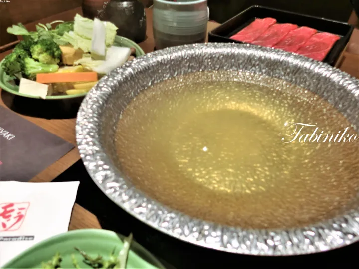 A simmering hot pot with clear broth, surrounded by plates of fresh vegetables and raw beef slices.
